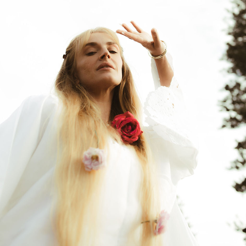 Woman with long blonde hair wearing a white dress with floral decorations, standing outdoors.